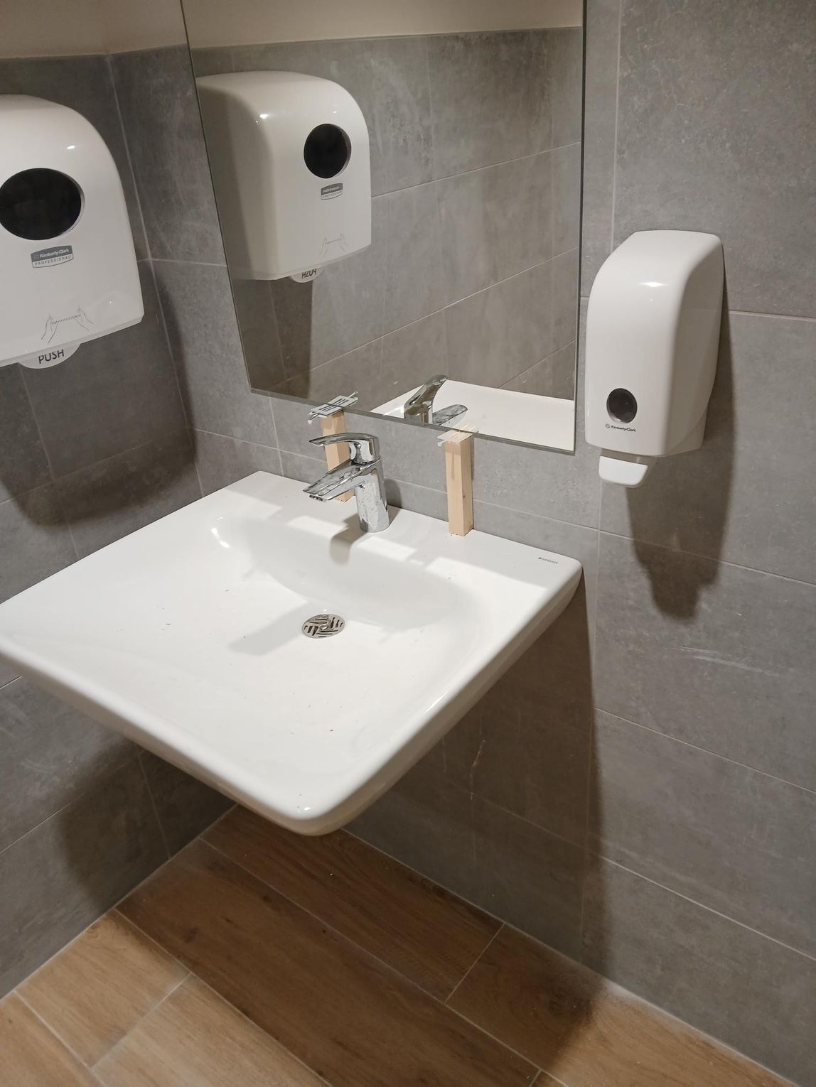 Lavabo blanc avec robinet, miroir et distributeurs de savon sur un mur carrelé.