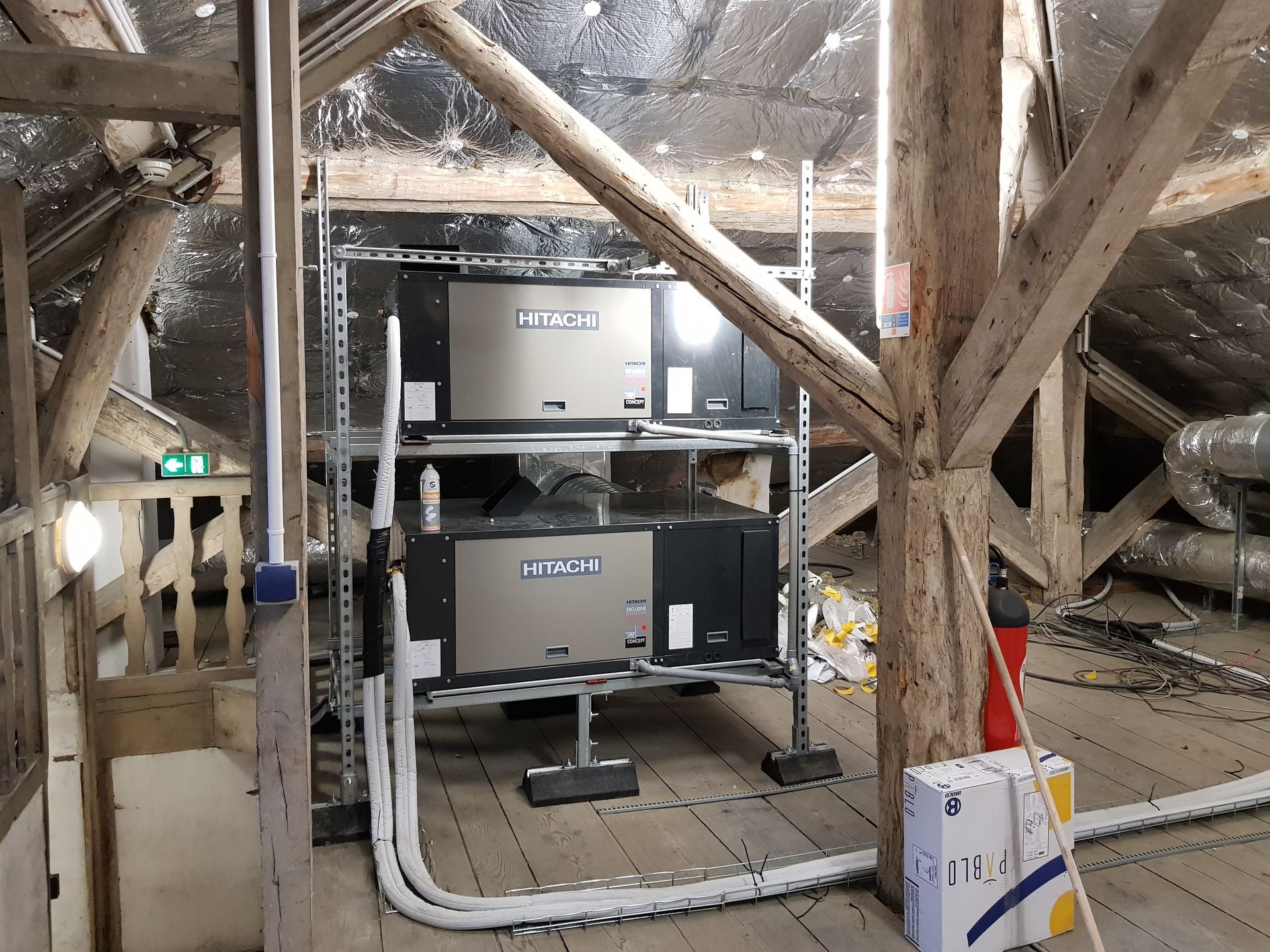 Unités de climatisation empilées sur une structure métallique dans un grenier aux poutres en bois et à l'isolation argentée.