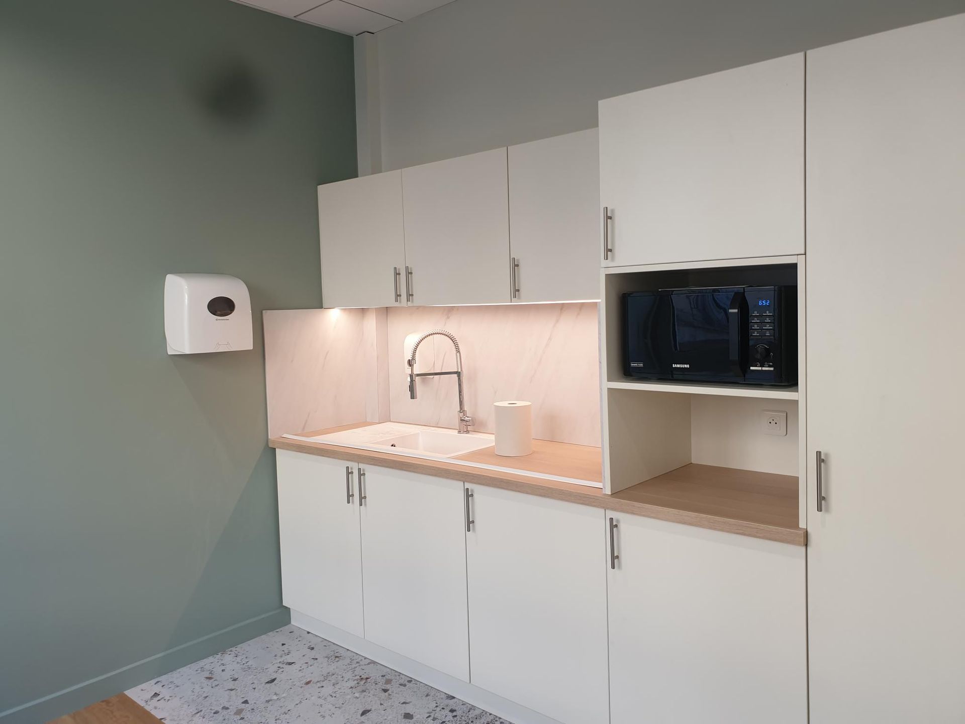 Une petite cuisine de bureau avec des placards blancs, un évier, un micro-ondes et un mur turquoise.