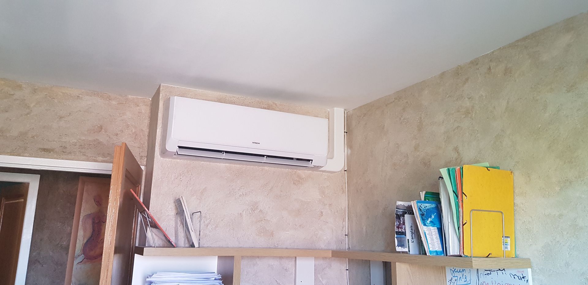 Un climatiseur est fixé sur un mur texturé, au-dessus d'une étagère. Des classeurs sont posés sur l'étagère.