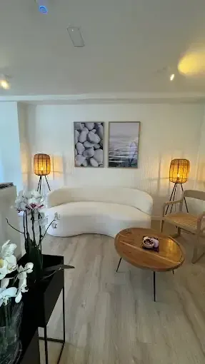 Acogedor salón blanco con sofá curvo, mesa de centro de madera, dos láminas enmarcadas y lámparas de pie.