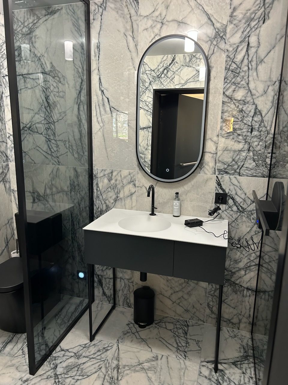 Salle de bains moderne avec murs en marbre, meuble-lavabo noir et miroir ovale. Une douche en verre et des toilettes sont également visibles.