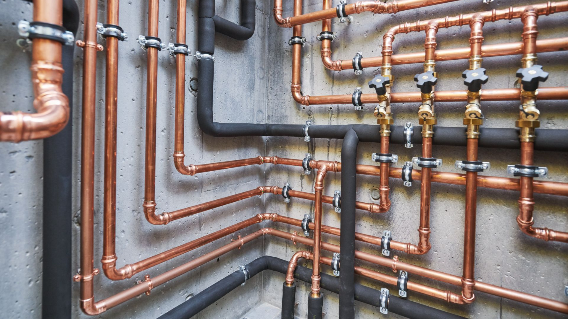 Des tuyaux en cuivre et en acier noir sur un mur en béton, formant un système de plomberie avec des vannes et des joints.