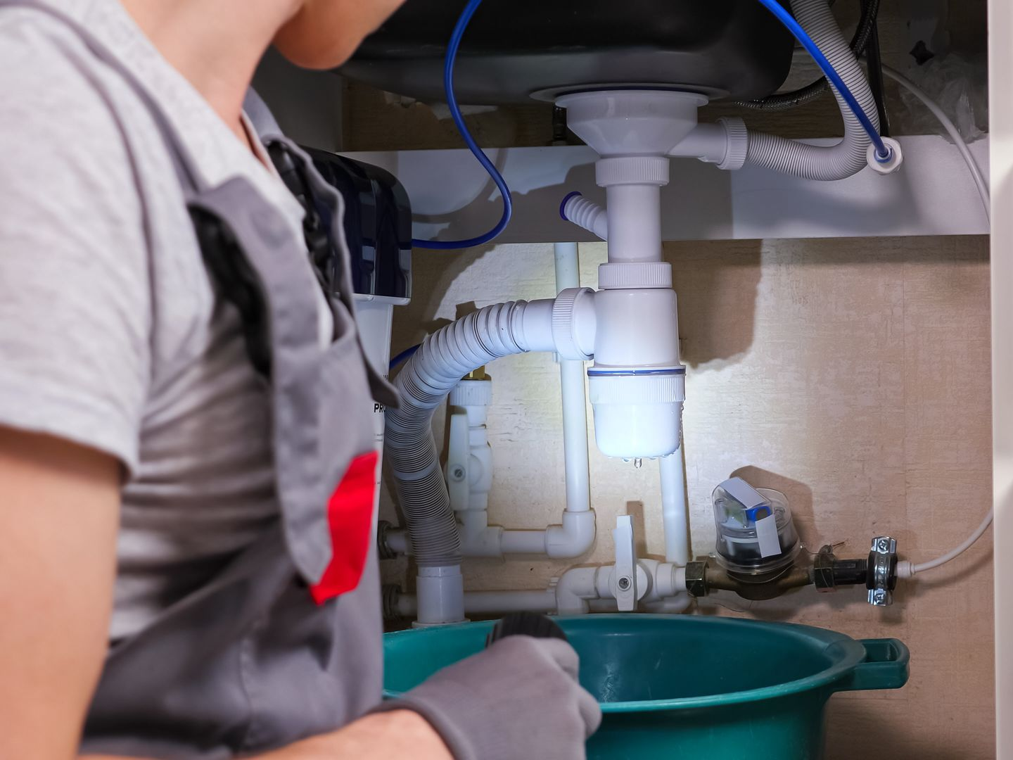 Un plombier travaille sur les tuyaux d'évier sous un lavabo, en tenant un récipient pour recueillir l'eau.