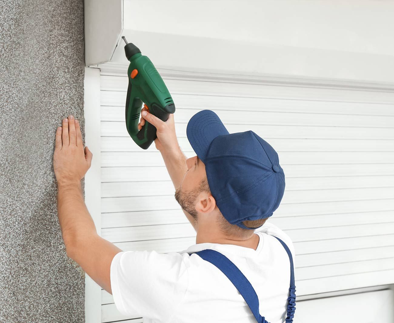 Persona con gorra azul y mono usando un taladro para instalar persianas blancas.