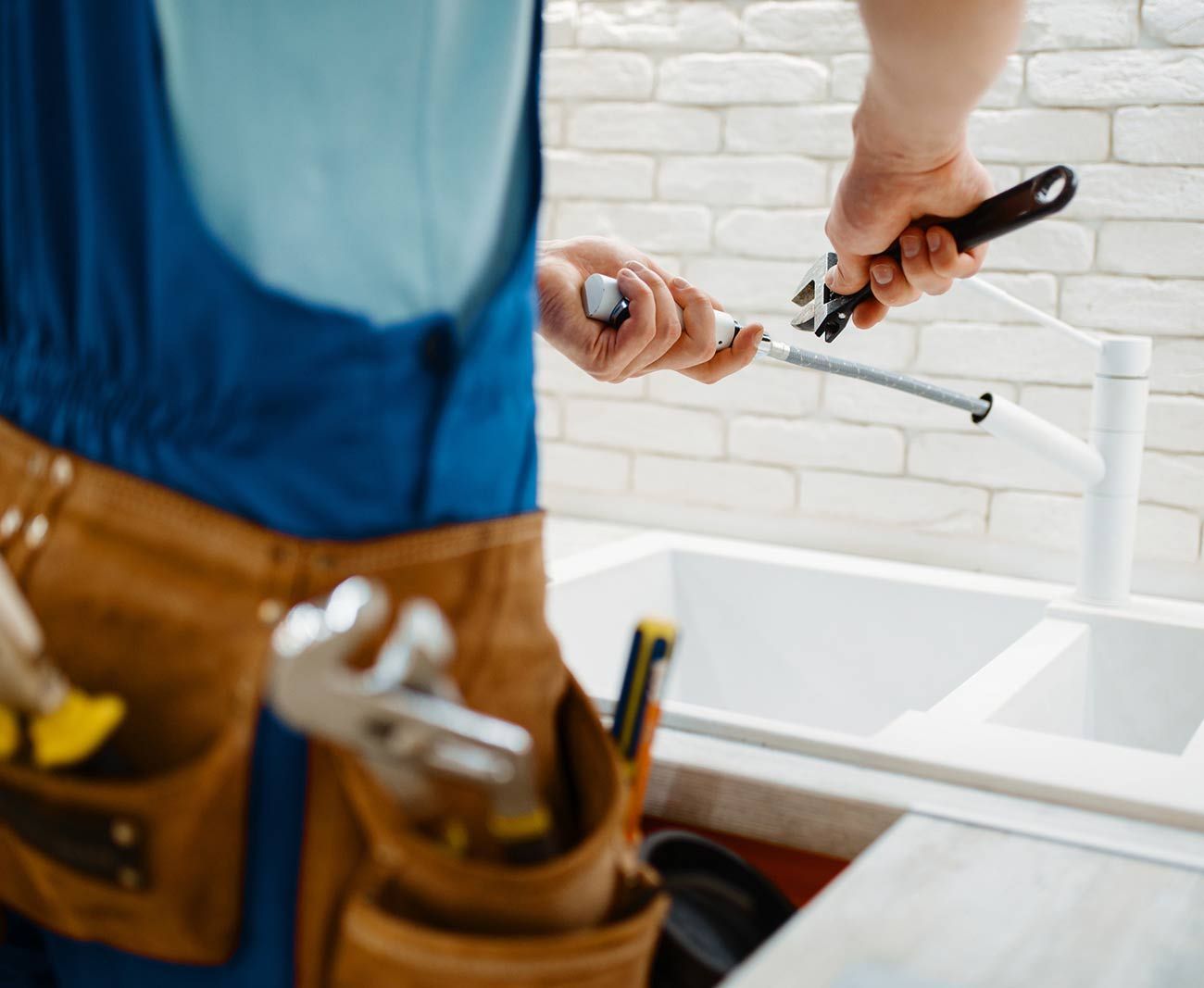 Un fontanero con mono azul usa una llave inglesa para apretar una tubería cerca de un fregadero blanco. Cinturón de herramientas visible.
