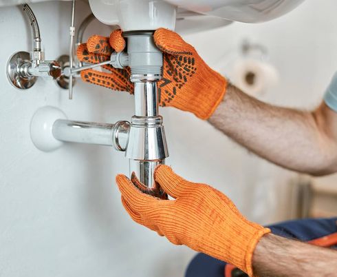 Unas manos enguantadas de naranja aprietan el tubo de desagüe cromado de un fregadero blanco.