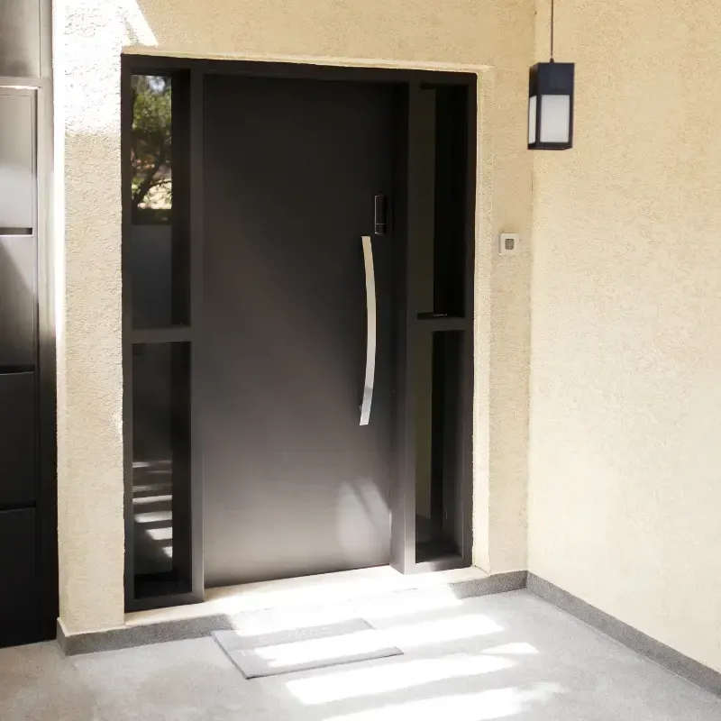 Puerta principal negra con paneles laterales, manija plateada y lámpara colgante en una pared de estuco color beige claro.