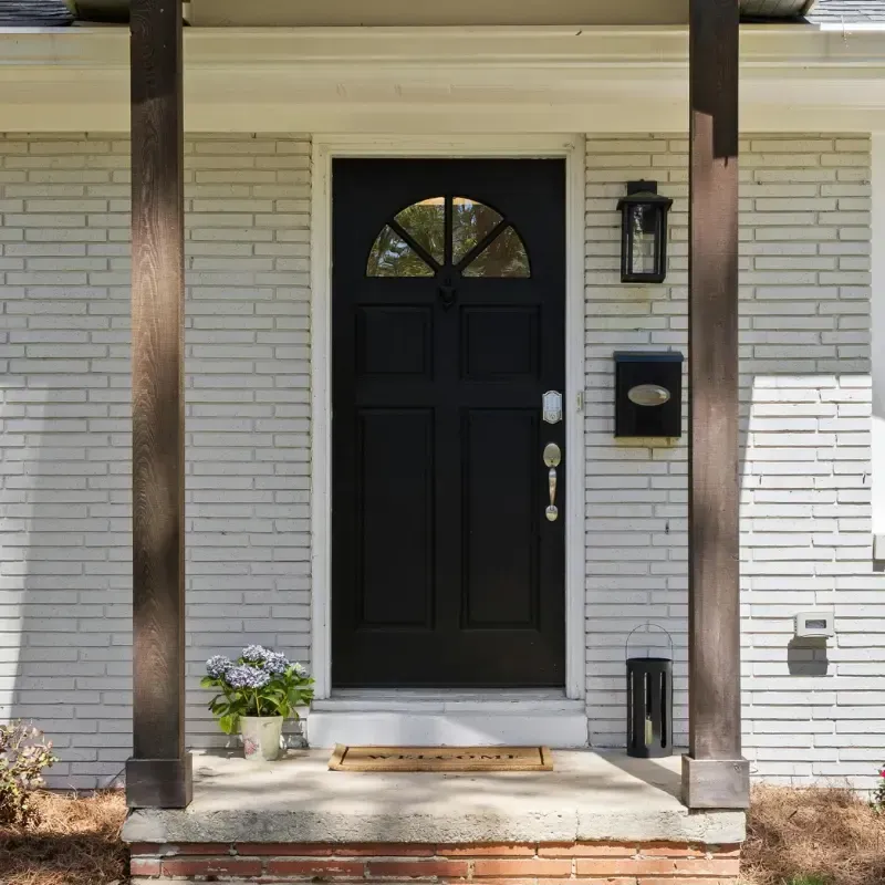 Puerta principal negra con cristal arqueado, rodeada de ladrillo blanco y columnas marrones.