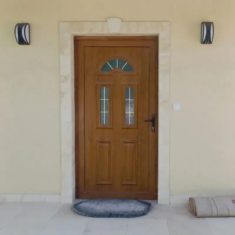 Puerta principal de madera con ventana arqueada, flanqueada por dos apliques negros, sobre una pared beige.
