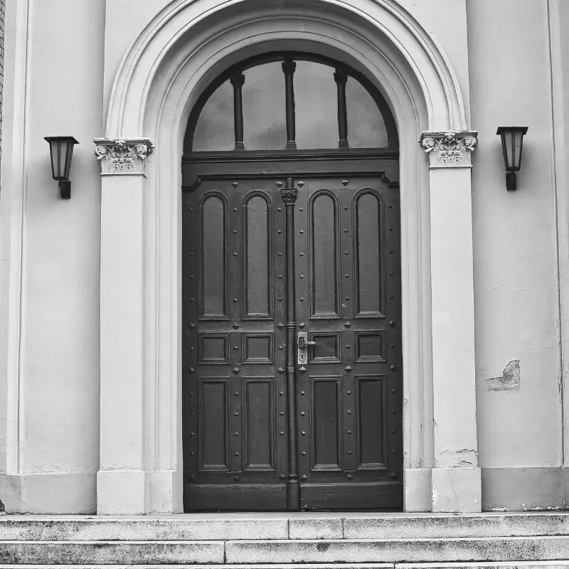 Fotografía en blanco y negro de una puerta doble de madera oscura con una ventana arqueada, flanqueada por pilares y faroles.