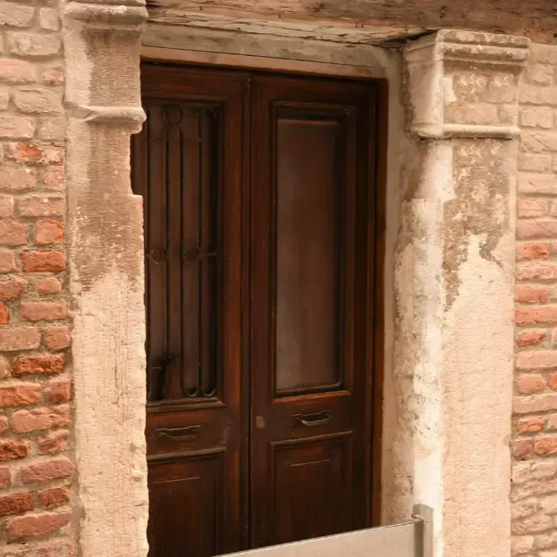 Puertas dobles de madera oscura en la entrada de un edificio de ladrillo desgastado, enmarcadas por piedra, con una ventana y una pequeña barandilla.