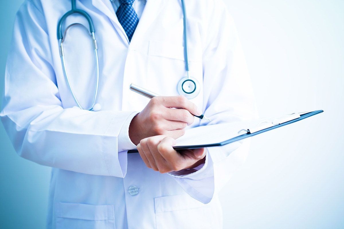 Médico con bata blanca escribiendo en una tabla con sujetapapeles y con un estetoscopio alrededor del cuello
