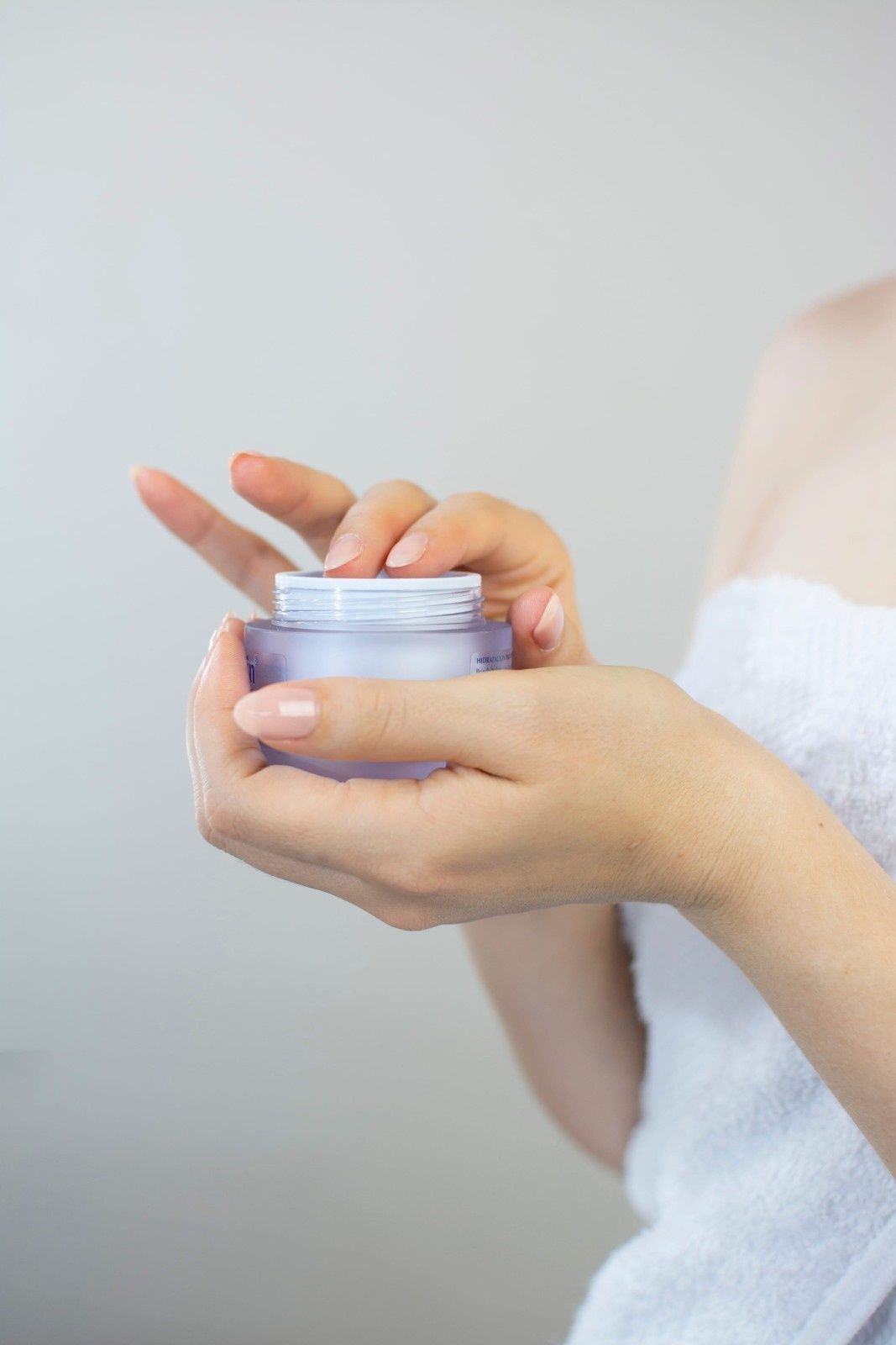 Una persona envuelta en una toalla blanca sostiene un pequeño frasco abierto de crema facial de color lila claro.