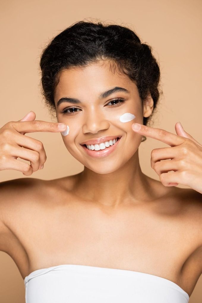 Una persona sonriente se aplica pequeños puntos de crema blanca en las mejillas sobre un fondo neutro liso.