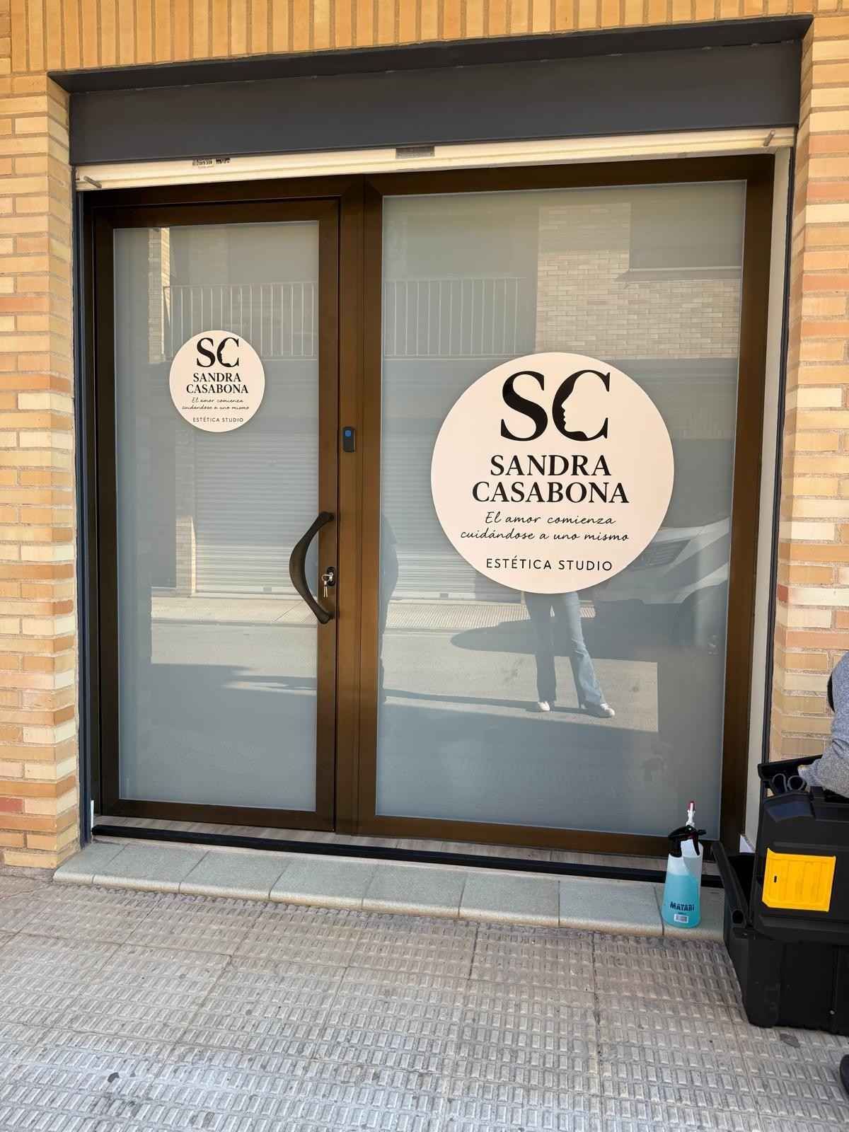 Local comercial con puertas de cristal y letreros circulares “SC Sandra Casabona” en un edificio de color beige.