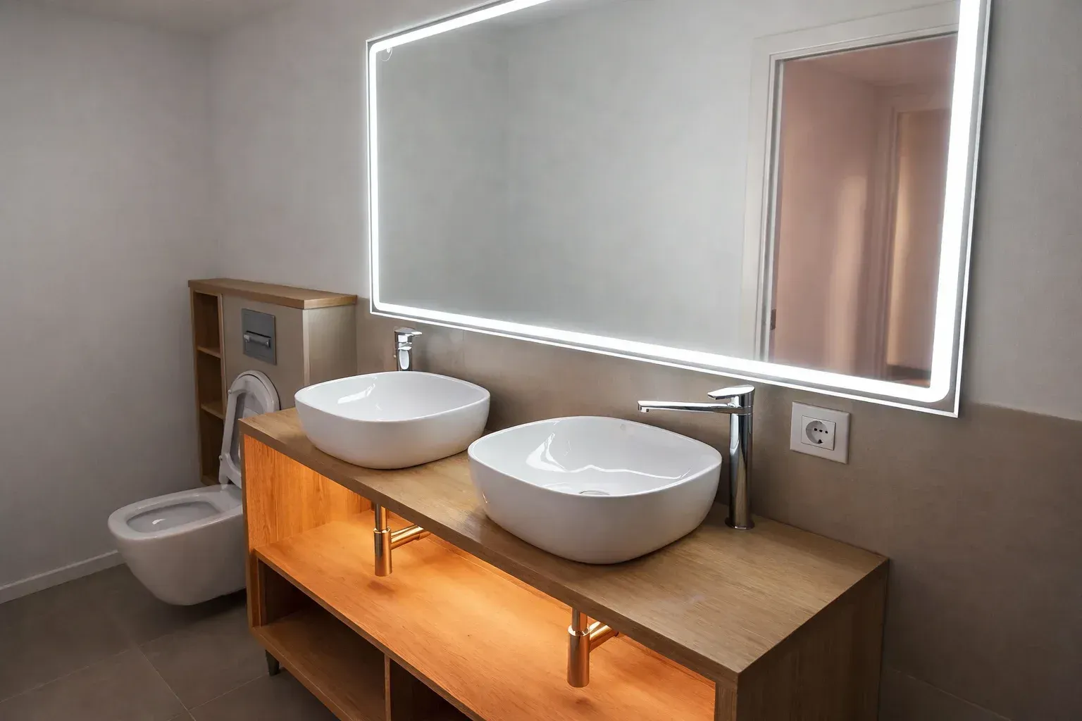 Baño moderno con dos lavabos blancos sobre un tocador de madera, un gran espejo iluminado y un inodoro montado en la pared.