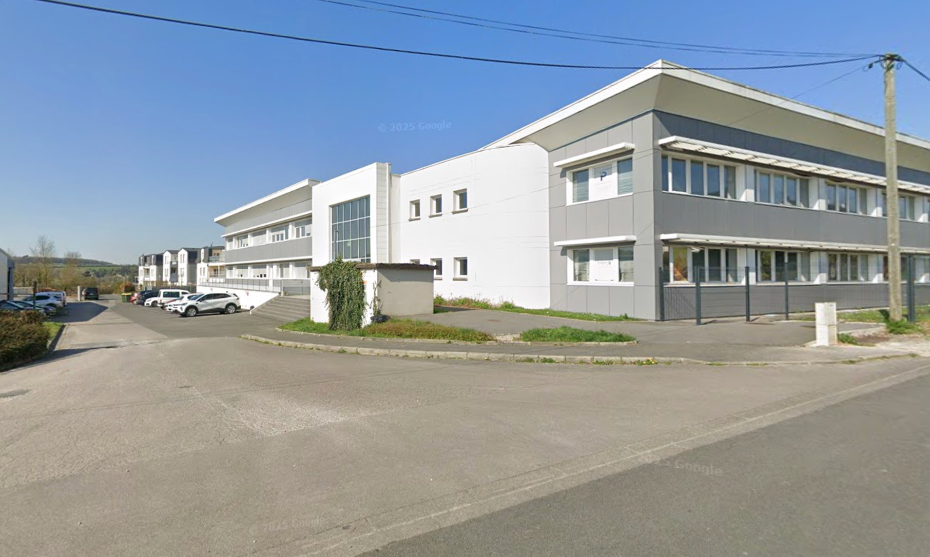 Bâtiment moderne de deux étages avec un revêtement gris, des touches de blanc et un petit rond-point. Ciel bleu au-dessus.