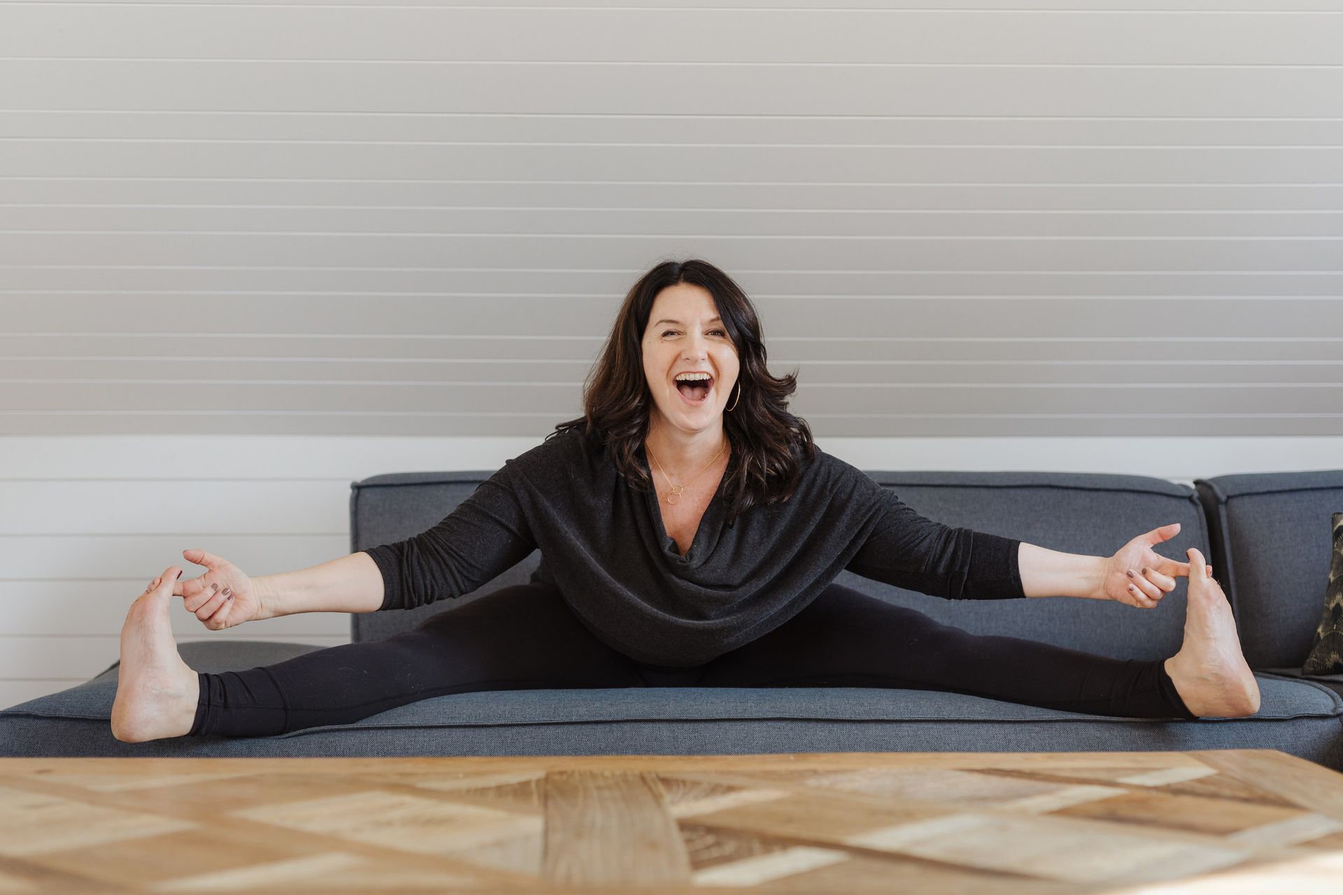 Eine Frau in schwarzer Sportkleidung macht lachend mit ausgestreckten Armen und Beinen den Spagat auf einem Sofa.