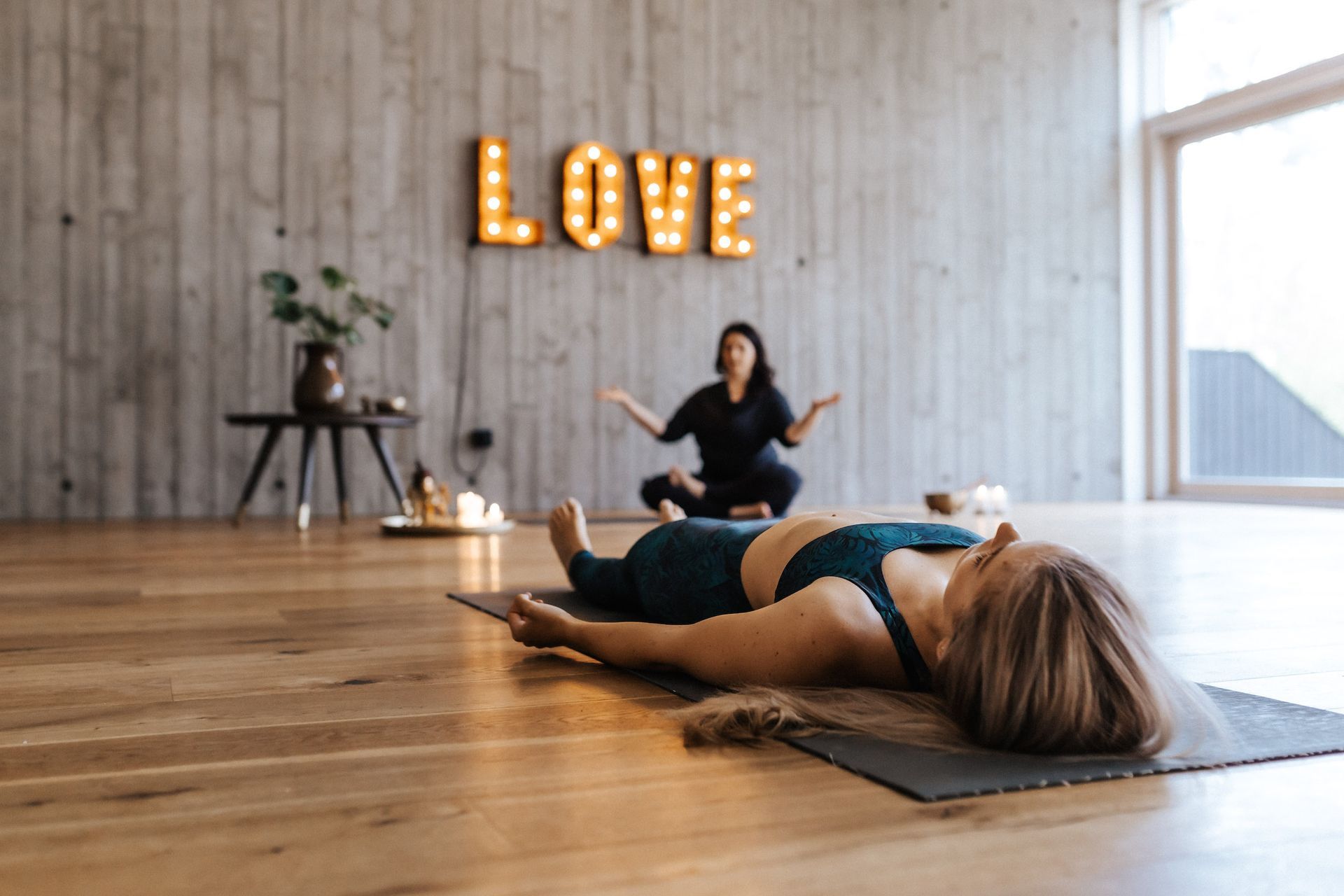 Yogalehrerin im Hintergrund, beleuchtetes „LOVE“-Schild an der Wand.