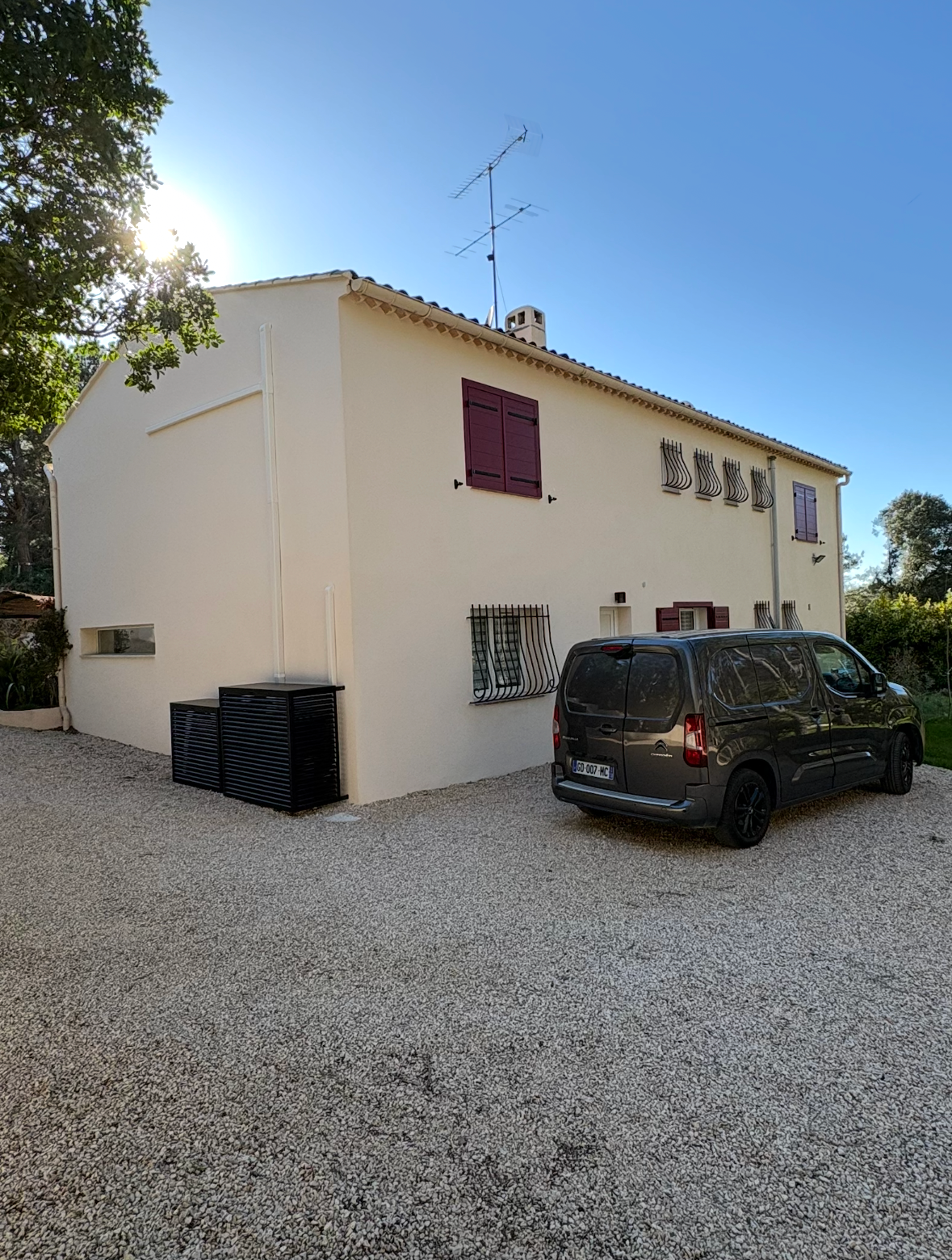 Deux unités de climatisation superposées sur un mur  beige, à côté d'une fenêtre à volets blancs et d'un buisson vert