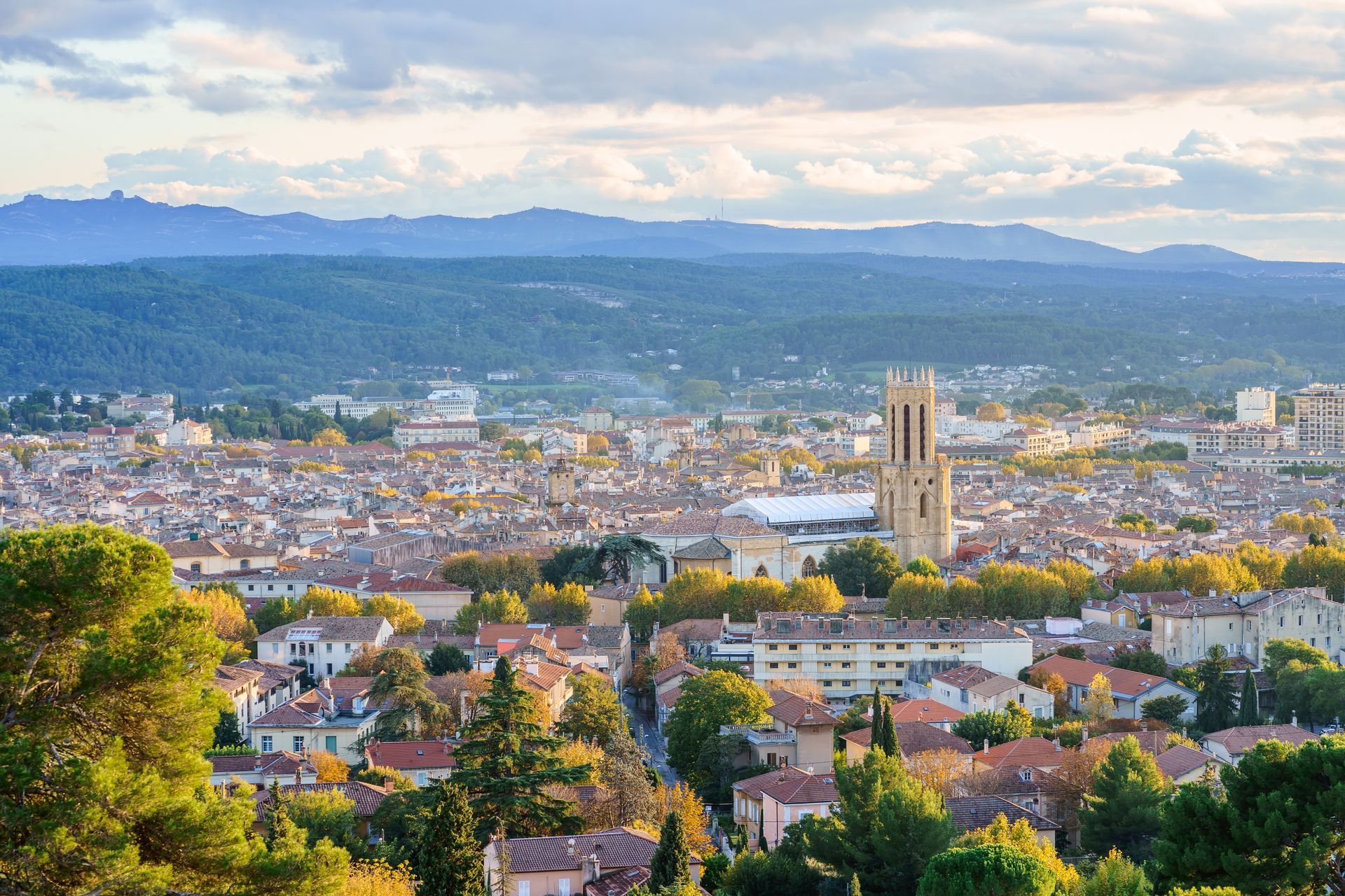 Vue d'Aix-en-Provence avec sa haute cathédrale et ses toits, et les montagnes en arrière-plan sous un ciel bleu.