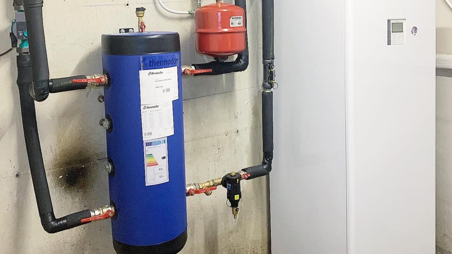 Chauffe-eau bleu et vase d'expansion rouge avec tuyaux noirs, paroi blanche et unité blanche haute.