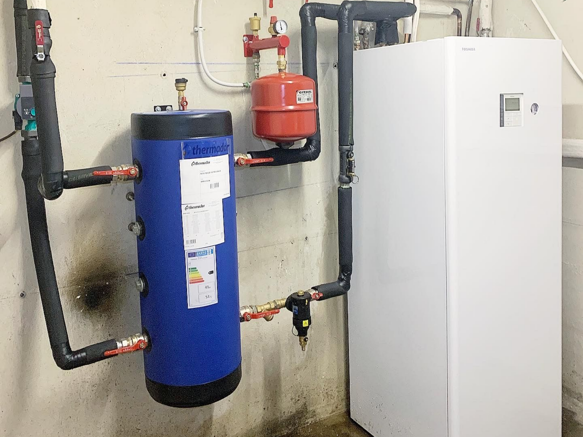 Composants bleus et blancs d'un système de chauffage fixés sur un mur blanc dans une buanderie.