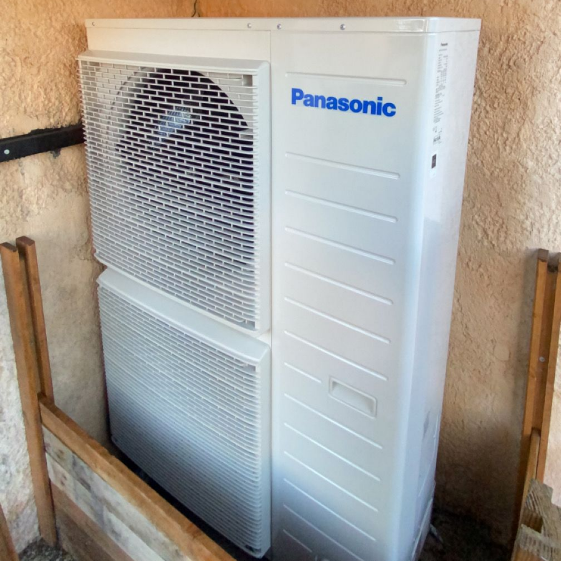 Unité de pompe à chaleur Panasonic extérieure, blanche avec logo bleu, à côté d'une clôture en bois.