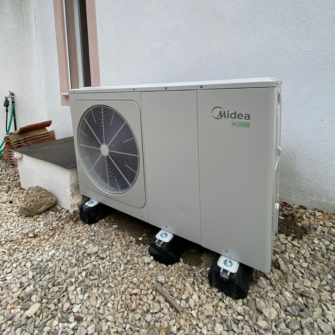 Une unité de pompe à chaleur Midea grise, installée à l'extérieur, repose sur des supports de montage noirs.