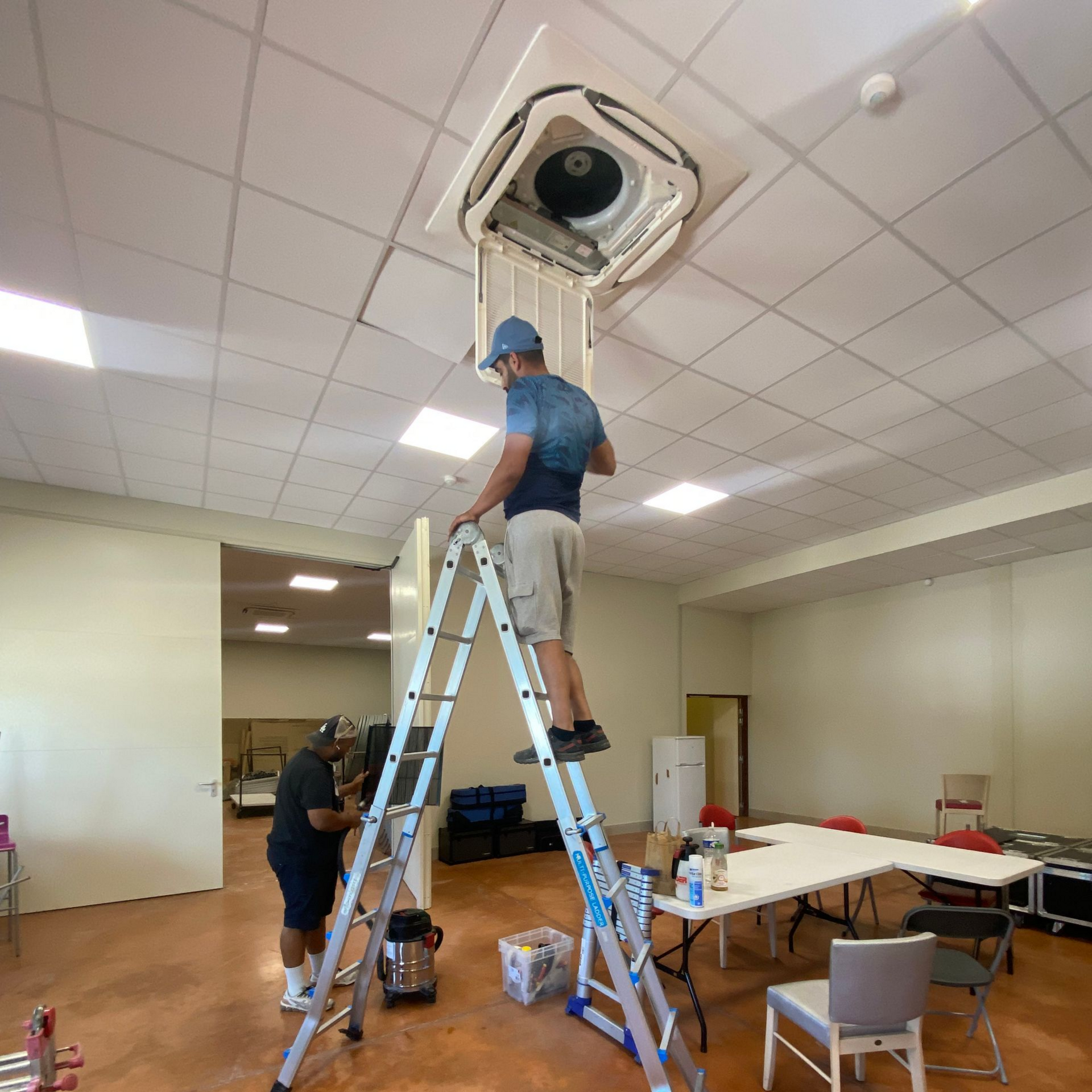 Deux personnes travaillent sur un climatiseur suspendu, à l'aide d'une échelle, à l'intérieur d'une pièce.