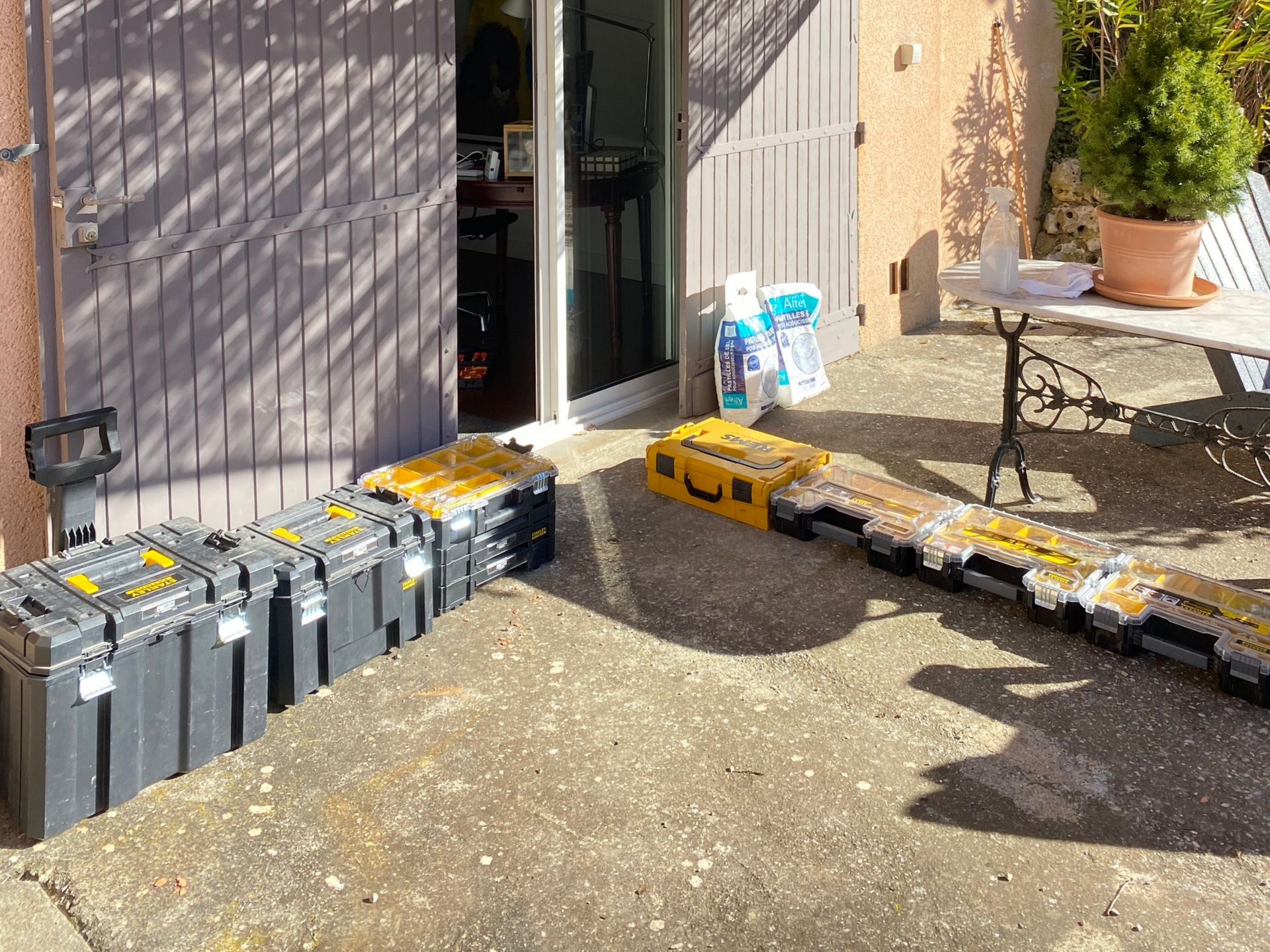 Plusieurs boîtes à outils alignées sur une terrasse en béton, à côté d'une porte, par une journée ensoleillée.