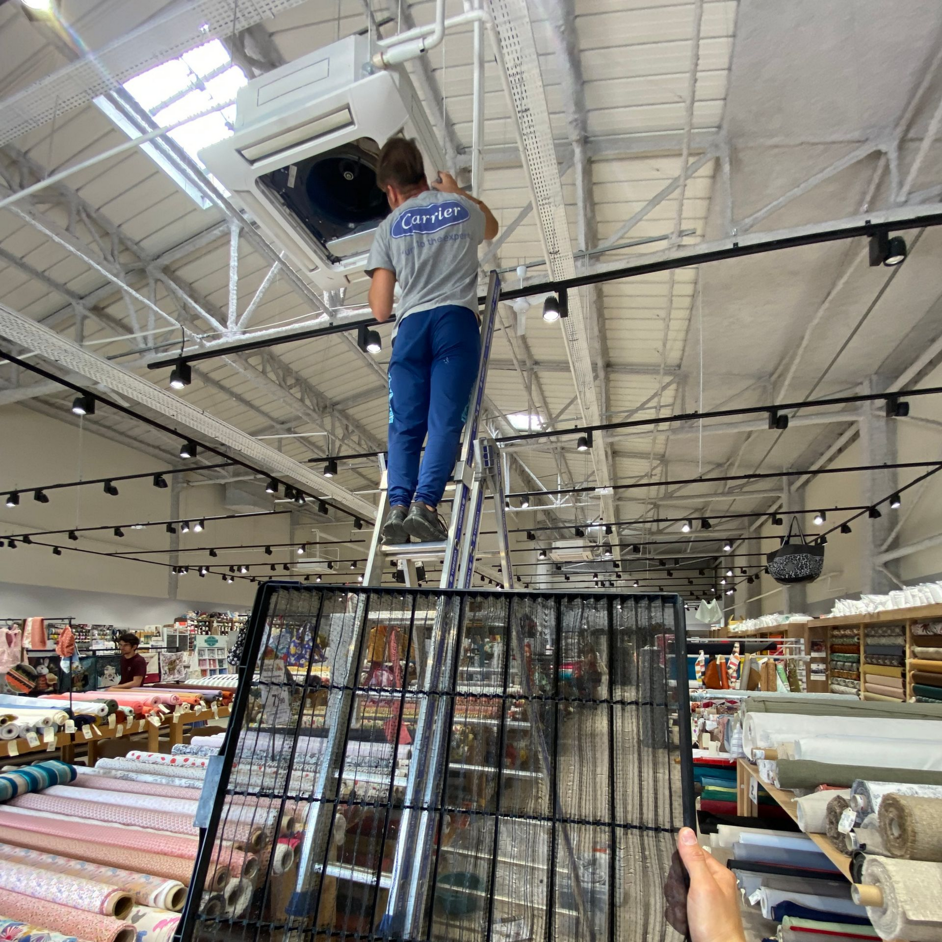 Une personne sur une échelle travaille sur un climatiseur fixé au plafond d'un magasin rempli de marchandises et éclairé.