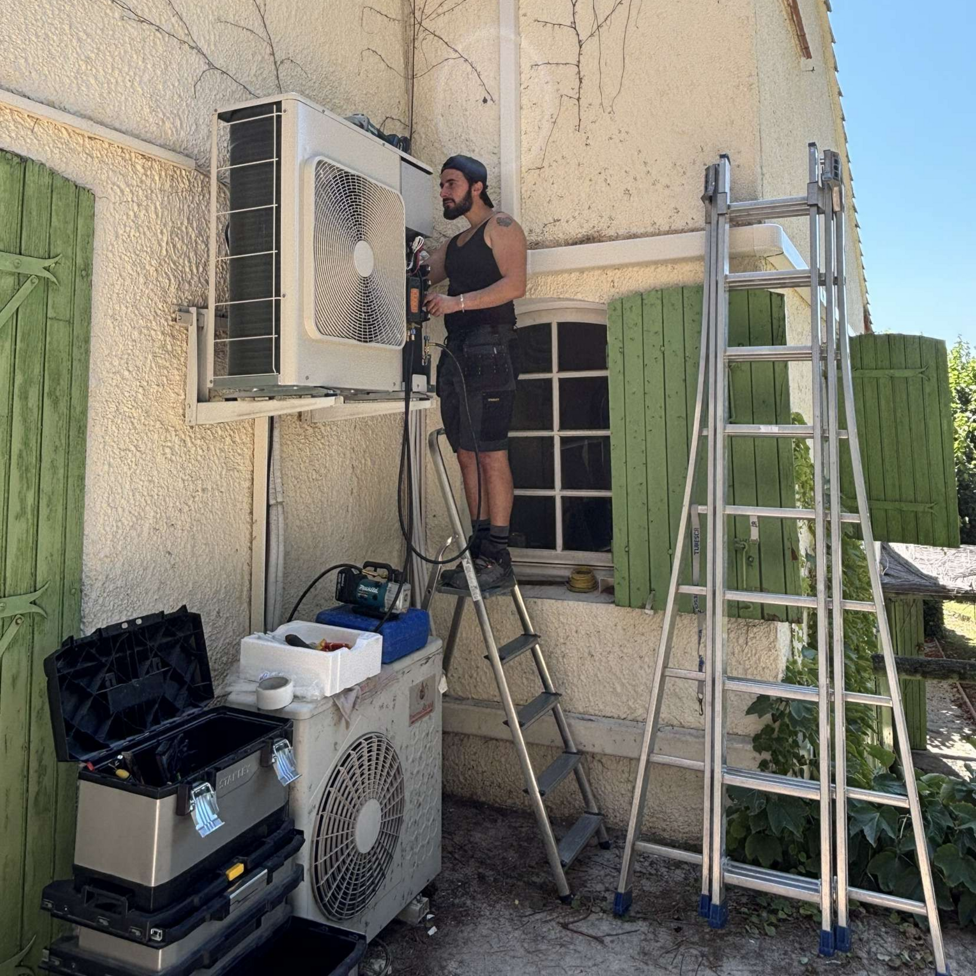 Loïc de LD-Energies installe un climatiseur extérieur sur une maison, à l'aide d'échelles. Les outils sont à proximité.