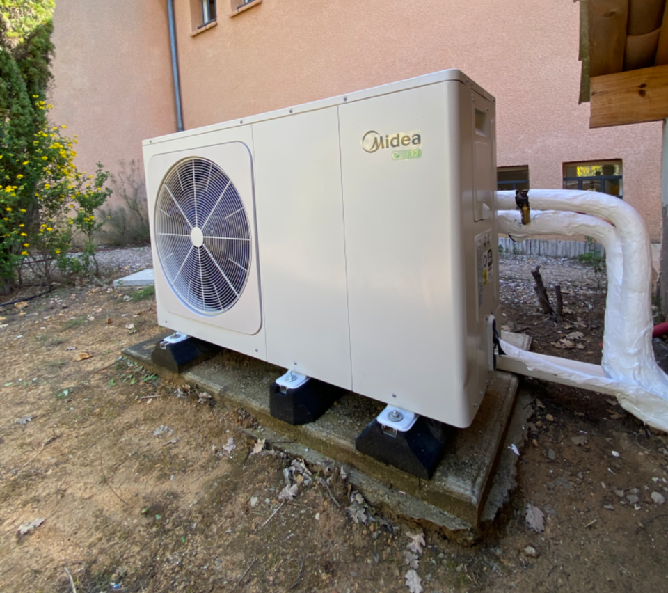 Pompe à chaleur Midea sur socle en béton, unité beige avec ventilateur, à l'extérieur d'un bâtiment, tuyauterie.