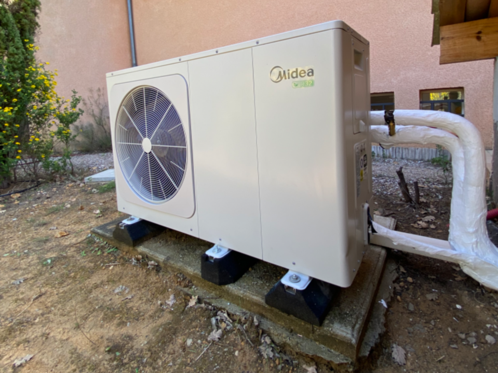 Pompe à chaleur air-air Midea sur socle en béton, couleur beige, avec tuyaux de protection, installation extérieure.