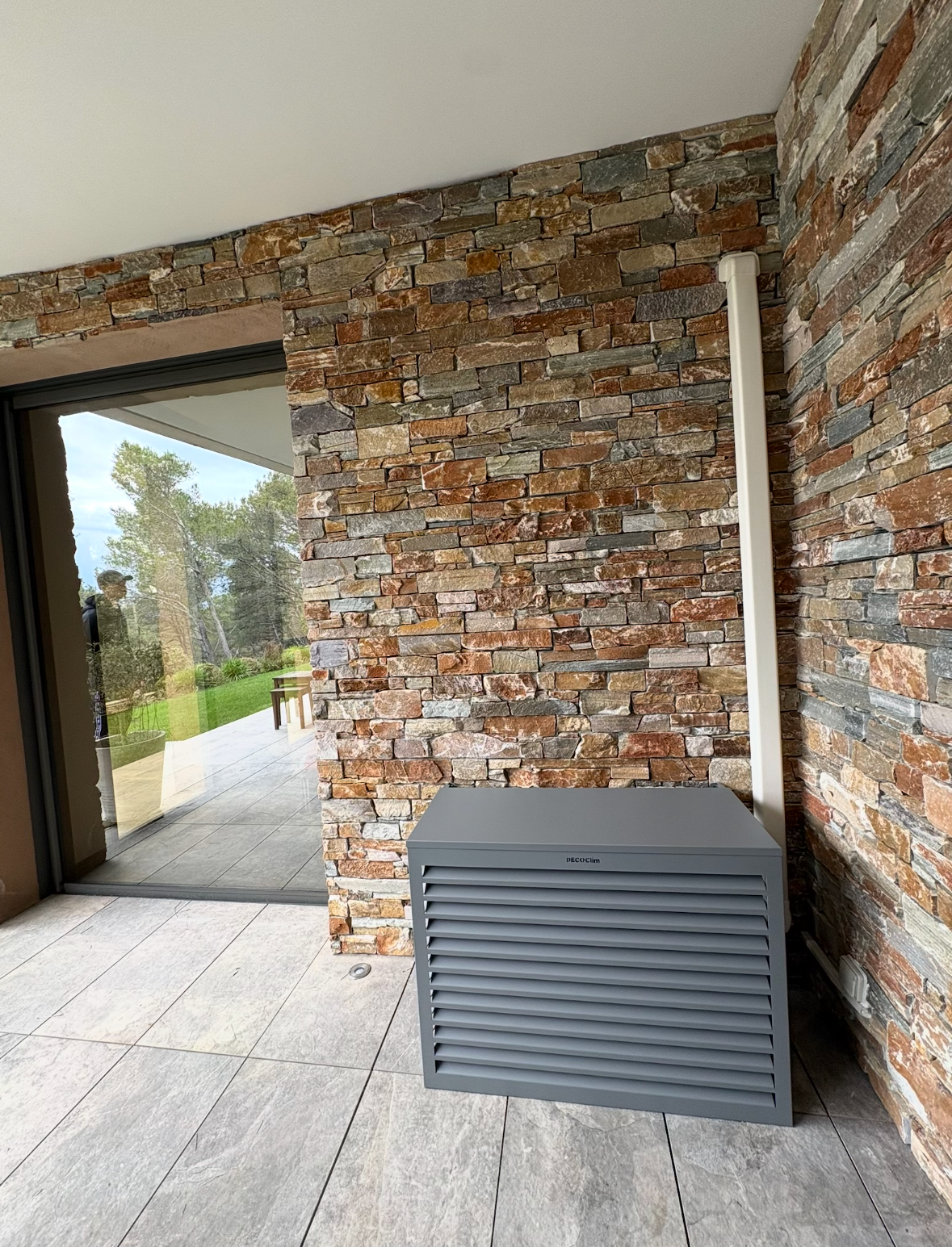 Un meuble rectangulaire gris à lattes est installé sur une terrasse carrelée, adossé à un mur de pierre rustique, à côté d'une porte vitrée offrant une vue