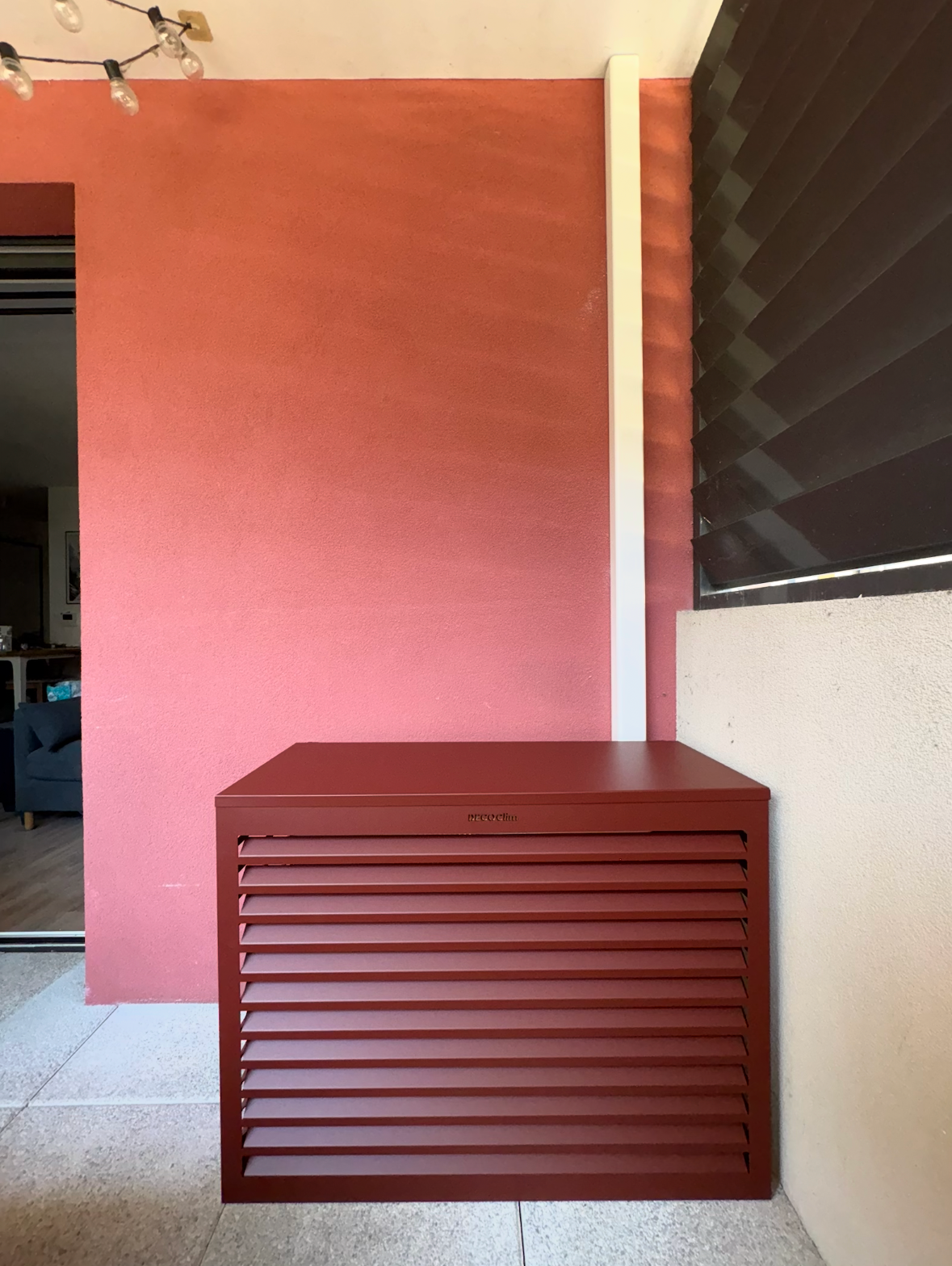 Un cache pompe à chaleur en métal rouge foncé adossé à un mur rouge assorti sur une terrasse, à côté d'un tuyau vertical blanc