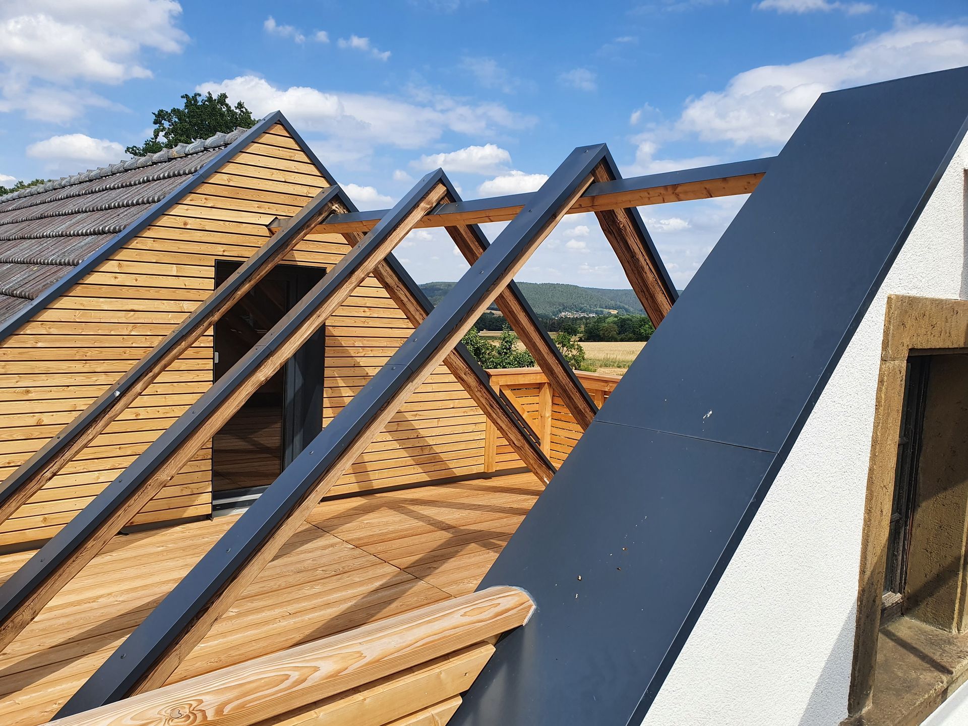 Im Bau befindliches Holzdach mit dunklem Metall und Balken, blauer Himmel im Hintergrund.