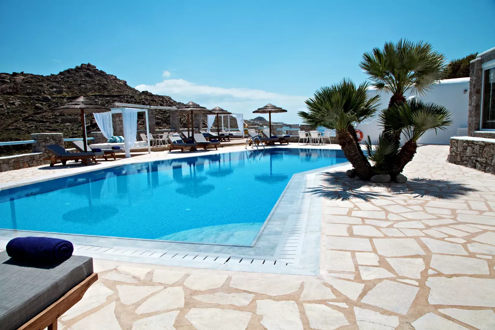 Sonnendurchfluteter Resort-Pool mit Liegestühlen, Sonnenschirmen und Steinterrasse neben felsigen Hügeln.