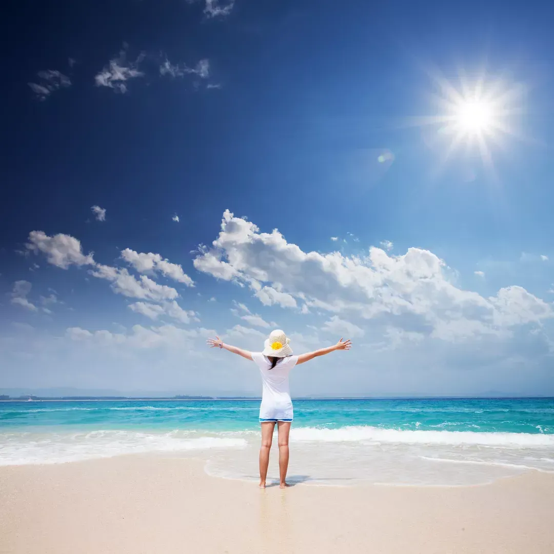 Eine Person in weißem Badeanzug und Sonnenhut steht an einem sonnigen Strand und hat die Arme zum Meer erhoben.