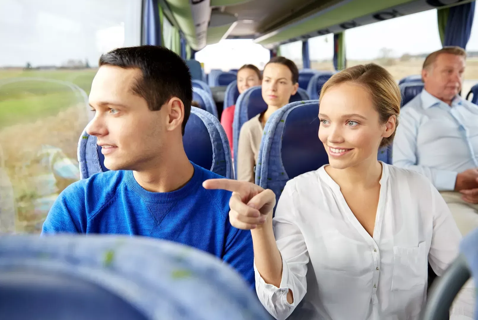 Fahrgäste unterhalten sich in einem Bus, eine lächelnde Frau zeigt zum Fenster.