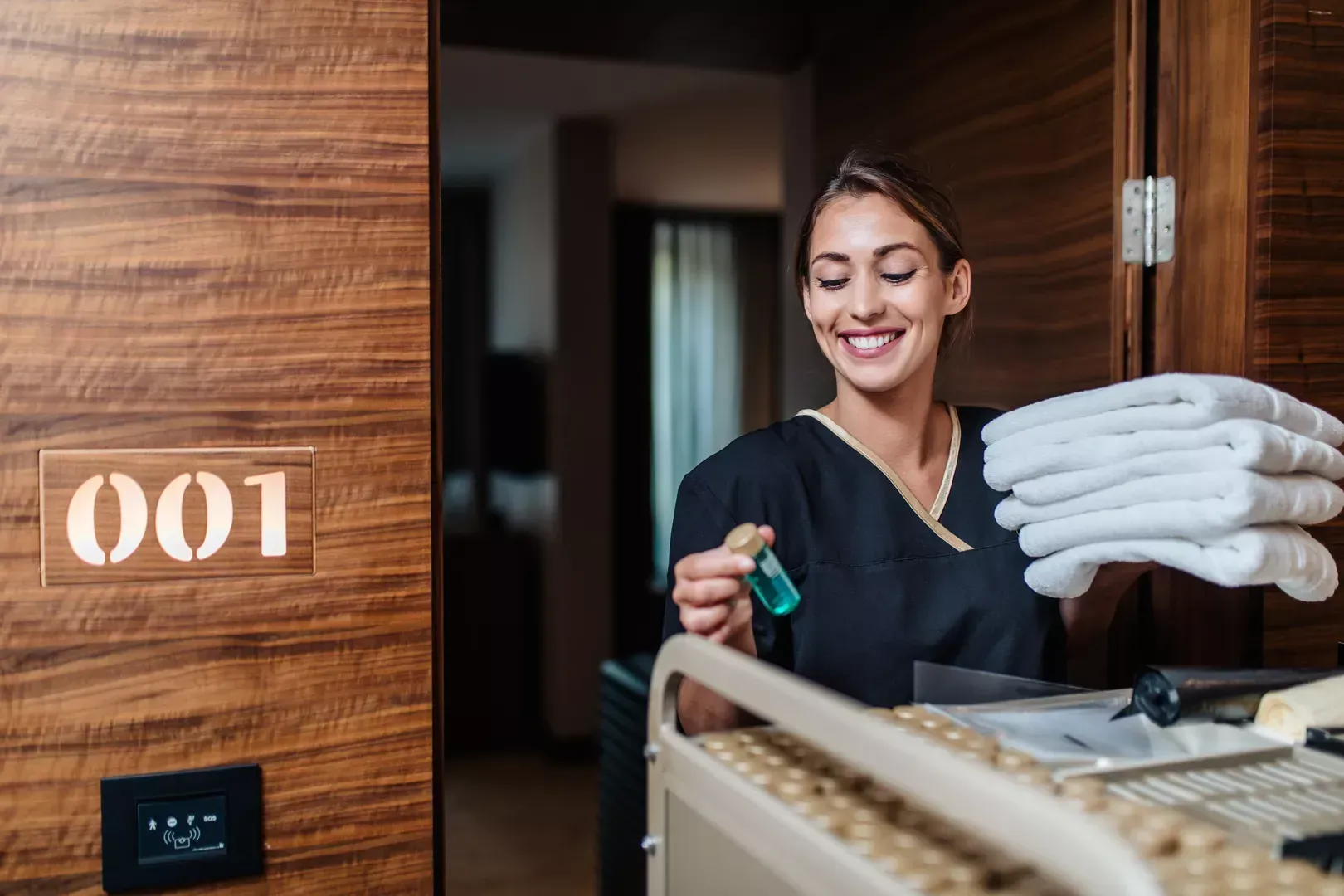 Eine lächelnde Zimmermädchen steht neben einem Stapel Handtücher in einem Hotelflur; Zimmer 001 ist an der Holzwand zu sehen.