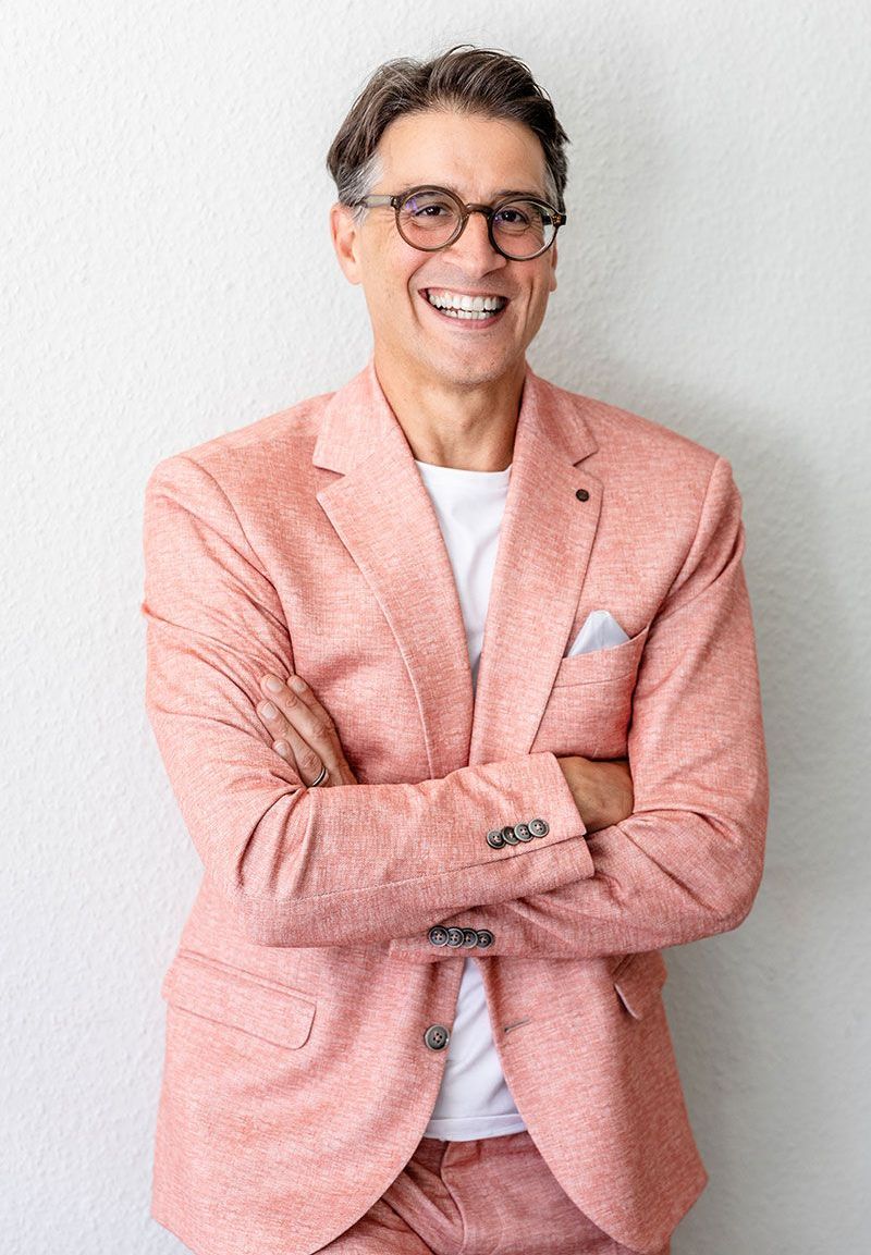 Ein Mann in einem rosa Anzug und mit Brille lächelt und verschränkt die Arme.