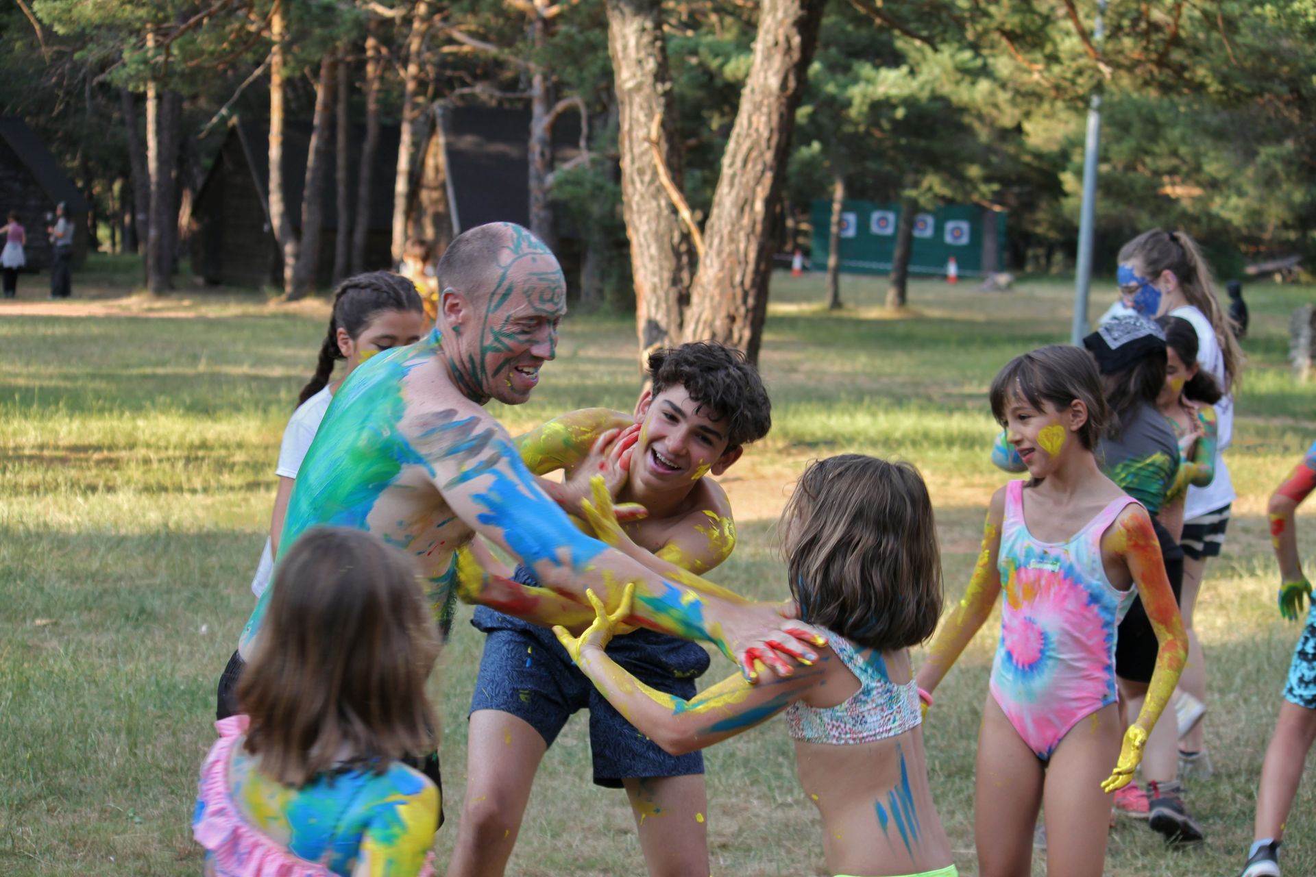Adultos y niños juegan con polvos de colores en un parque, sonriendo y extendiendo los brazos unos hacia otros.