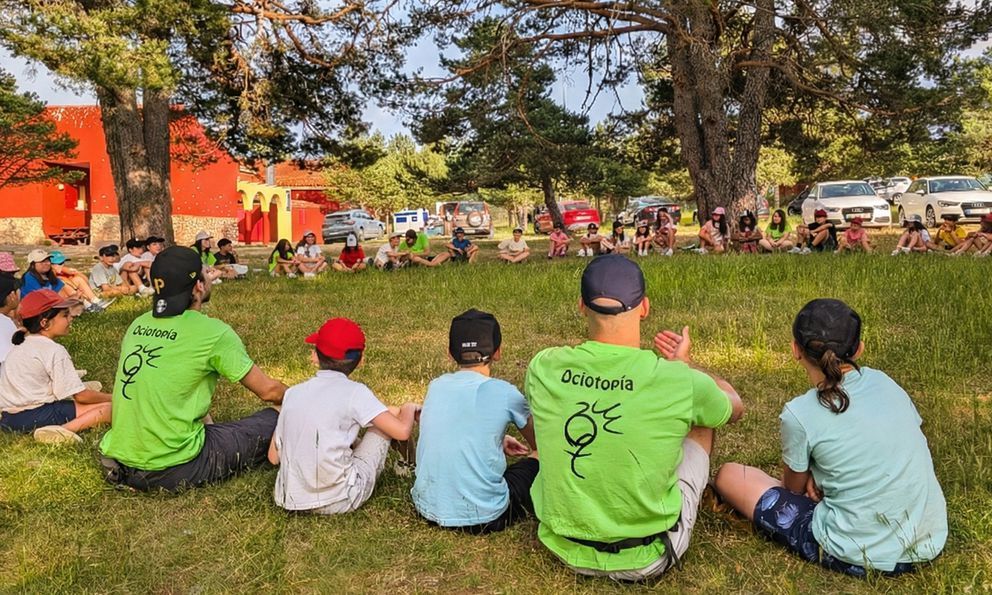 Gente sentada en el césped de un parque, observando un evento al aire libre cerca de árboles y carpas.