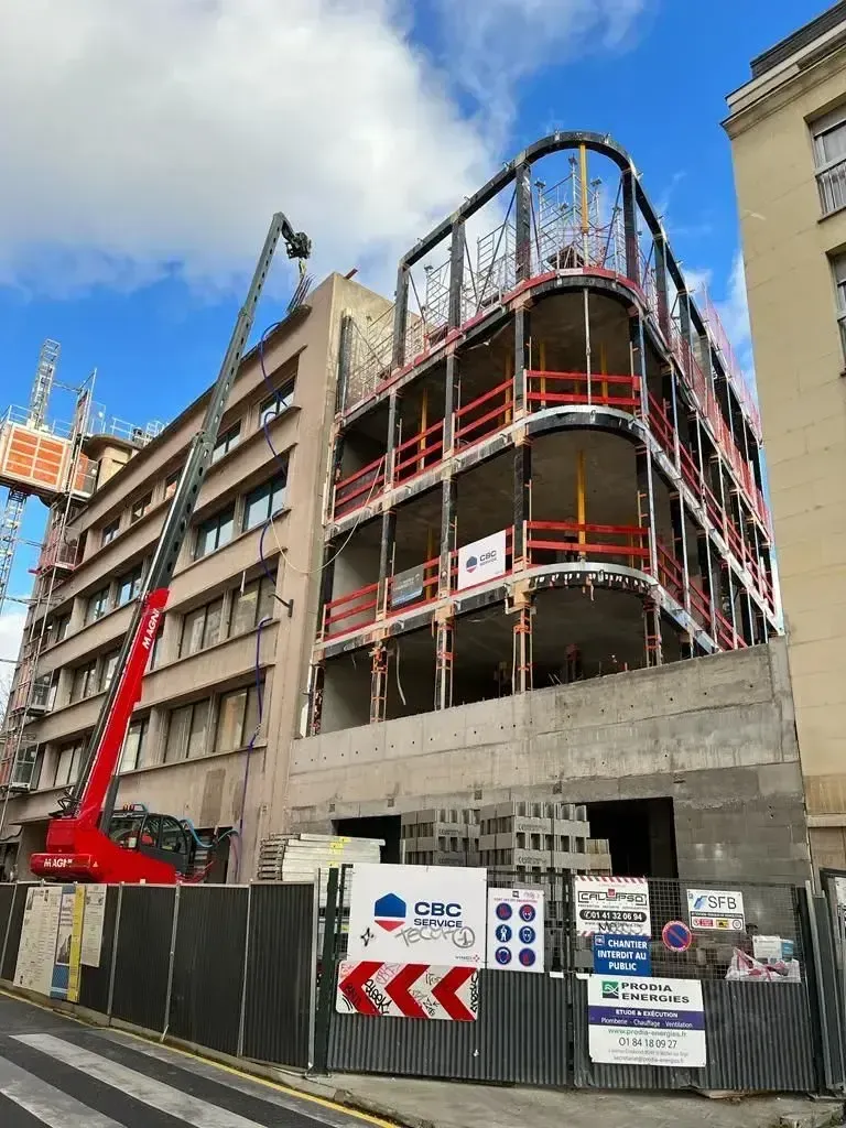 Chantier avec une nacelle élévatrice rouge à bras articulé près d’un bâtiment en rénovation, derrière une clôture noire et des panneaux de signalisation