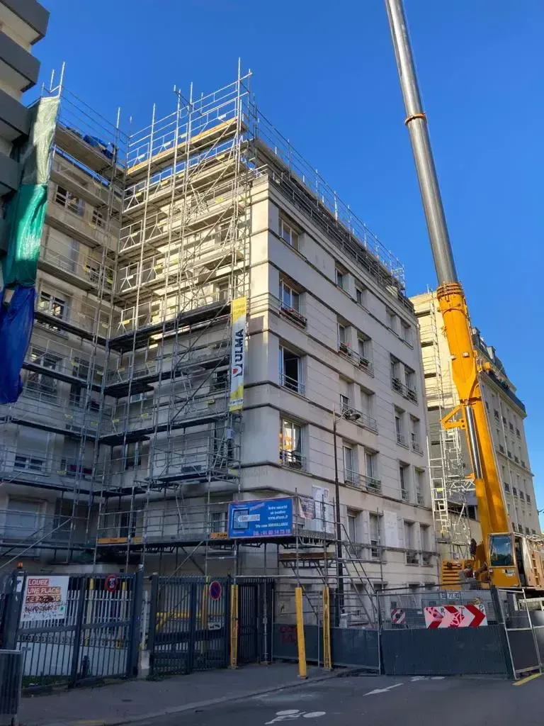 Angle d’un immeuble en construction dans une rue de la ville, avec des barrières de chantier et une grue jaune sous un ciel bleu
