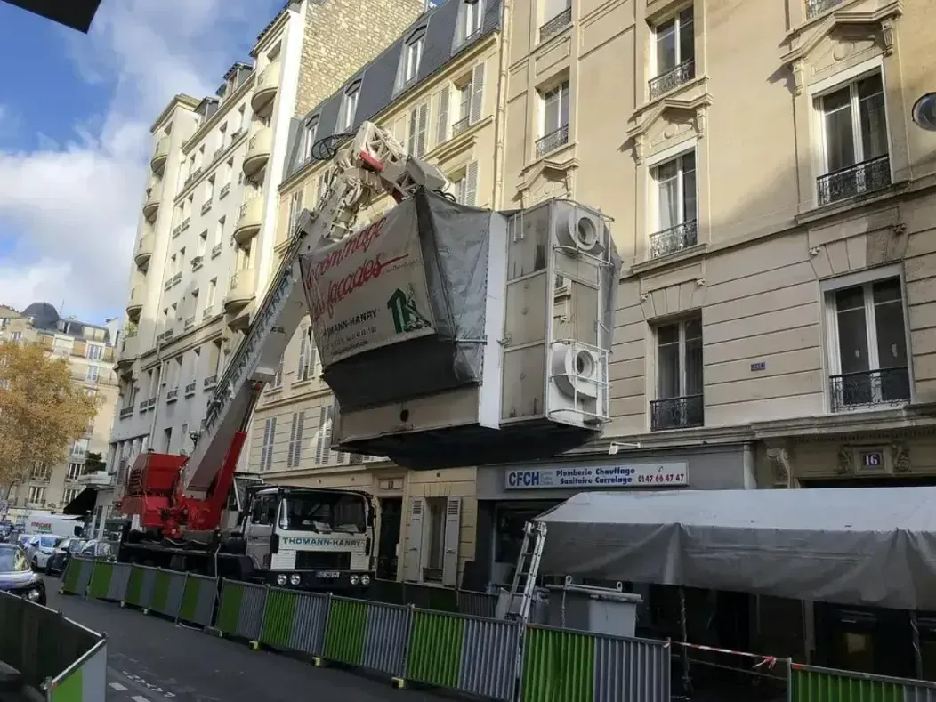 Plateforme de travaux urbains soulevant une grande caisse à côté d’un immeuble de style parisien et d’une terrasse de café