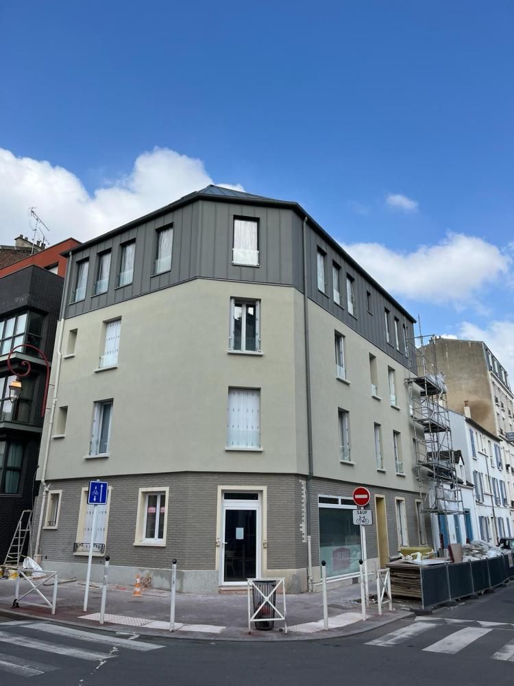 Immeuble d’angle gris dans une rue sous un ciel bleu, avec des panneaux de signalisation et des barrières